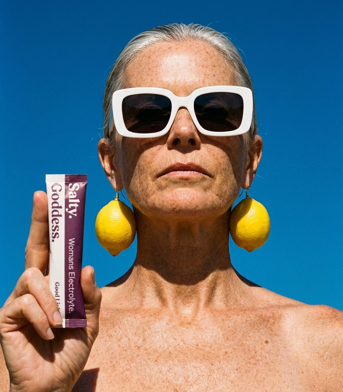 A woman in white sunglasses and lemon earrings holding a Salty Goddess stick pack against a blue sky